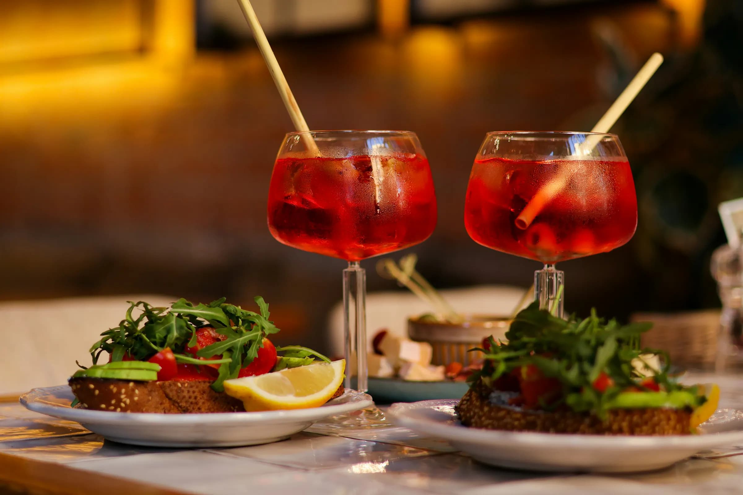 Due spritz con cannuccia e bruschette su un tavolo del bar, con luci calde sullo sfondo